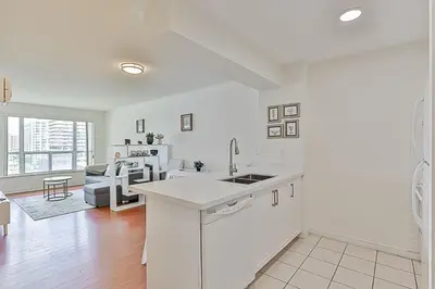 a white kitchen with a sink and a living room at Cozy 1BR Condo with Balcony and Parking in Toronto