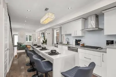 a white kitchen with a long counter and chairs at Entire Unit - Steps From History - 5 Mins To Beaches in Toronto