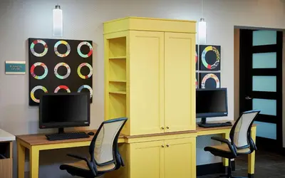 a yellow cabinet with two monitors on a desk with chairs at Home2 Suites By Hilton Brantford in Brantford