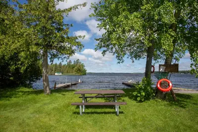 a picnic table in the grass next to a lake at Fun Place with a Sandy Beach for Swimming, Boating & Fishing, Featuring 1 House and 3 Cabins in Fenelon Falls